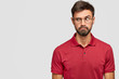 Candid shot of puzzled Caucasian male with dark stubble looks suspiciously aside, raises eyebrows, wears red t shirt, notices something on blank space. People and facial expressions concept.