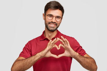 Wall Mural - Friendly attractive bearded male makes heart gesture, smiles pleasantly, wears spectacles and red t shirt, expresses love to girlfriend, isolated over white background. Body language concept