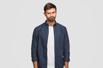 Wall Mural - Gloomy unshaven guy has irritated expression, raises eyebrows and purses lips, doesn`t like something, expresses his discontent, wears white t shirt and dark blue shirt, poses in studio alone