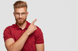 Studio shot of attractive young ginger male with thick beard, points at upper right corner, has self confident facial expression, wears red t shirt, isolated over white background with copy space