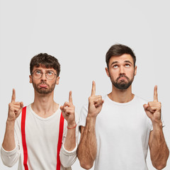 Wall Mural - Photo of sullen men with dark hair, dressed in white clothes, points upwards, have displeased facial expressions, indicate at copy space for your advertisement or promotional text. Two male companions