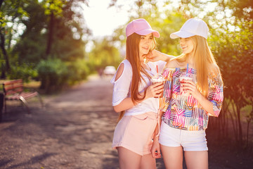 Confident two women in summer outfits and caps standing, walking together with refreshing drinks in countryside