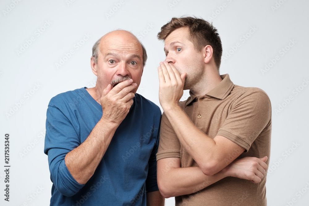 Young caucasian son telling secret to his senior father. Senior man in shock opening his mouth. - obrazy, fototapety, plakaty 
