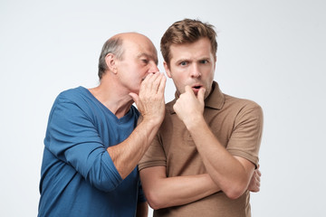 senior man sharing secret or whispering gossips into his son ear. telling family secret concept