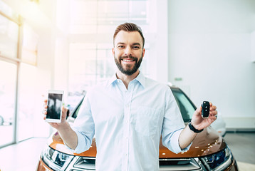 Wall Mural - Apps for buying car easy. Young bearded man shows car key and phone with blank screen on camera in dealership