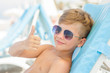 © Ermolaev Alexandr - the boy on a sunny beach lies on a sun lounger in sunglasses and showing thumbs up