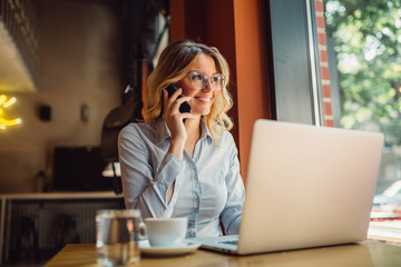 portrait of young business woman with glasses sitting in cafe in front of her laptop and talking on 