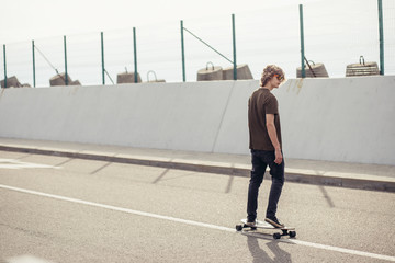 Wall Mural - Young curly long haired man riding on longboard on empty road