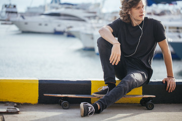 Wall Mural - skater boy sitting on his longboard on pier and looking at yacht, take break after riding