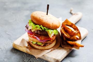 Tasty grilled beef burger with bacon and french fries served on a rustic wooden board on dark slate background