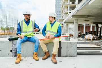 young man in hardhat and waistcoat holding cup of hot beverage and gesturing while telling story to 