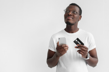Closeup photo of young African male in eyeglasses pictured isolated on gray background thinking and dreaming about pleasant plans with credit card and smartphone in hands, ready to purchase in web