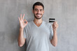 © Damir Khabirov - Portrait of young smiling man in t shirt, holding credit card and showing okay sign, standing against gray textured wall