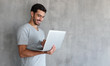 © Damir Khabirov - Indoor portrait of young man in t shirt standing against textured wall with copy space for ads, holding laptop and watching media with happy smile, sharing web content