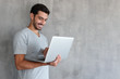 © Damir Khabirov - Young handsome man standing against textured gray wall with copy space for ads, holding laptop and watching media with happy smile, sharing web content
