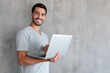 © Damir Khabirov - Indoor portrait of young man in t shirt standing against textured wall with copy space for ads, holding laptop and looking at camera with happy smile