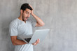 © Damir Khabirov - Portrait of handsome young man with mouth opened, in gray t shirt, holding silver laptop, looking surprised and confused, standing against  textured wall