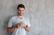 © Damir Khabirov - Young smiling man wearing wireless earphones and smart watches, standing against gray textured wall with copy space, looking at screen of smartphone