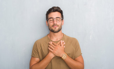Handsome young man over grey grunge wall wearing glasses smiling with hands on chest with closed eyes and grateful gesture on face. Health concept.