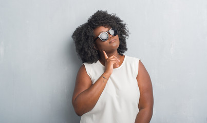 Poster - Young african american plus size woman over grey grunge wall wearing fashion sunglasses with hand on chin thinking about question, pensive expression. Smiling with thoughtful face. Doubt concept.