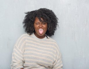 Canvas Print - Young african american plus size woman over grey grunge wall wearing a sweater sticking tongue out happy with funny expression. Emotion concept.