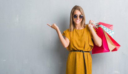 Wall Mural - Beautiful young woman over grunge grey wall holding shopping bags on sales very happy pointing with hand and finger to the side