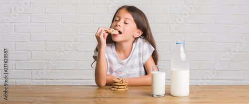 Fototapeta Young hispanic kid sitting on the table drinking milk and eating chocolate cooky with a confident expression on smart face thinking serious