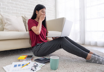 Poster - Asian woman freelance yawn while working on laptop with paperwork sitting one floor and leaning sofa at home,work at home concept.