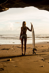 Young surfer girl with surf board pose in cave at beach. Garbage and plastic on sand. Concept of ecological problem and disaster. People water sport adventure camp and extreme swim on summer vacation