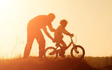 Silhouette Of Father Teaching Little Daughter To Ride A Bike At Meadow During Sunset. Caring Father Teaches Her Daughter To Ride Bicycle