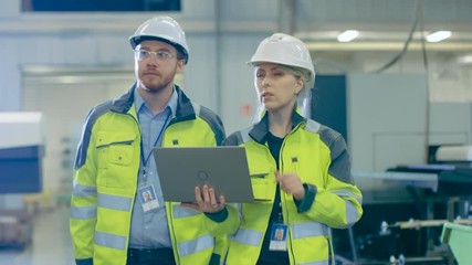 Poster - Male and Female Mechanical Engineers Talking, while Walking Through Industrial Factory, Male Holds Laptop. 