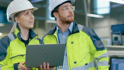 Poster - Male and Female Industrial Engineers Wearing safety,  Work on a Manufacturing Plant, They Discuss Project while Using Laptop.In the Background Industrial Facility.