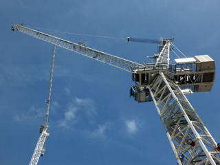 a perspective view of two tall white construction cranes on a building site against a blue sky with clouds