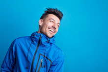 Portrait Of A Young Laughing Man In A Studio With Anorak On A Blue Background.