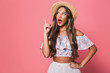 Portrait of excited pretty woman 20s wearing straw hat screaming and pointing finger upward, isolated over pink background in studio