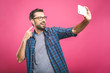 © denis_vermenko - I love selfie! Handsome young man in shirt holding camera and making selfie and smiling while standing against pink background. Listening music with headphones.