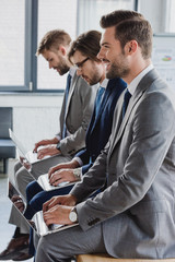 Wall Mural - side view of handsome young businessmen in suits sitting and using laptops in office