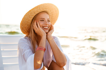 Sticker - Portrait of european cheerful woman 20s in straw hat smiling, while sitting in lounge chair at seaside during summer morning