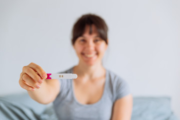 young happy woman sitting on bed looking on positive pregnancy test. blurred background