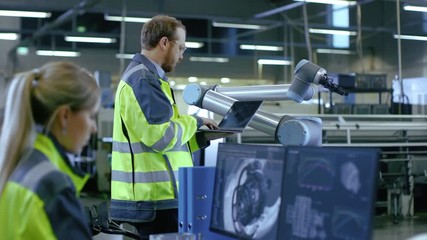 Poster - At the Factory: Male Automation Engineer Uses Laptop for Programming Robotic Arm while Female Mechanical Engineer Designs 3D Engine on Her Personal Computer.