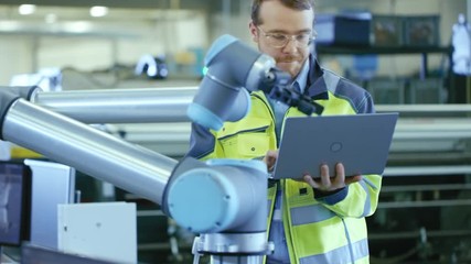 Poster - At the Factory: Automation Engineer Uses Laptop for Programming Robotic Arm. New Era in Automatic Manufacturing Industry.