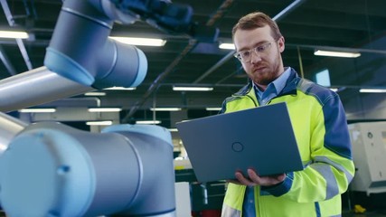 Poster - Low Angle Shot At the Factory: Automation Engineer Uses Laptop for Programming and Testing Robotic Arm. New Era in Automatic Manufacturing Industry.