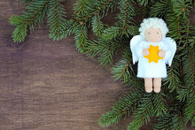 White Felt Christmas Angel With Yellow Star In Hands To Fresh Natural Branches Of Christmas Tree Spruce On Wooden Background