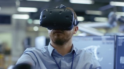 Poster - Portrait Shot of the Industrial Engineer Wearing Virtual Reality Headset and Using Controllers, ready to Work. In the Background Manufacturing Plant and Monitors.