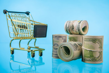 Money and a shopping cart. Money is twisted. Money is wrapped in an elastic band. Shopping cart of gold color. Expenses for purchases.