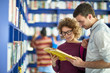 © pressmaster - Positive young students in casual clothing standing at blue shelf and reading book together while finding necessary information in library