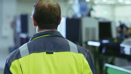 Poster - Following Back View Shot of the Industrial Engineer Wearing Protective Clothing Puts on Hard Hat and Walks Through Modern Manufacturing Facility with Automatic Machinery Working in Background.