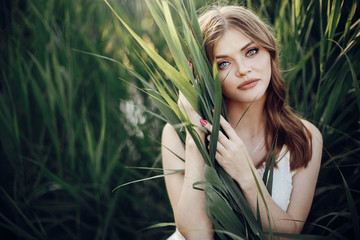 beautiful boho girl embracing in grass at sunset light near lake. attractive sensual young woman with amazing eyes and lips posing in white dress in green cane. save environment