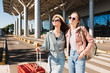Two pretty smiling girls in sunglasses joyfully looking aside with red suitcase and backpack near airport