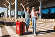 Two cheerful female friends in sunglasses happily looking in camera raising hands up with red suitcase and backpack on shoulder near airport